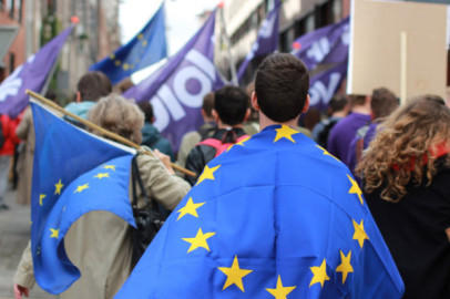 A man in the foreground wearing a European Flag. More European flags are seen in the background with people also waving Volt flags.