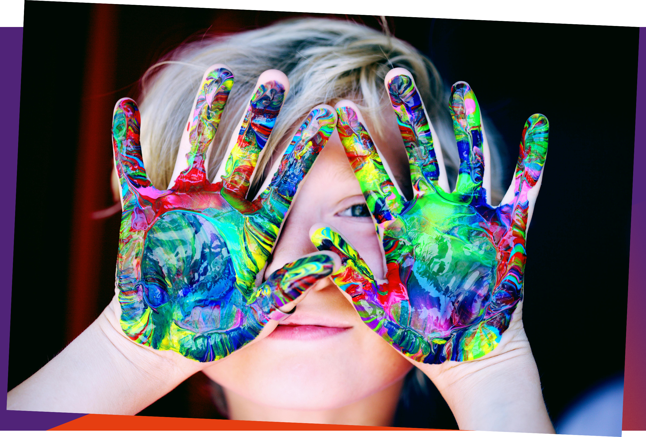 A blonde child holds two hands with the palms facing forward in front of her face. The palms of the hands are painted in all the colours of the rainbow.