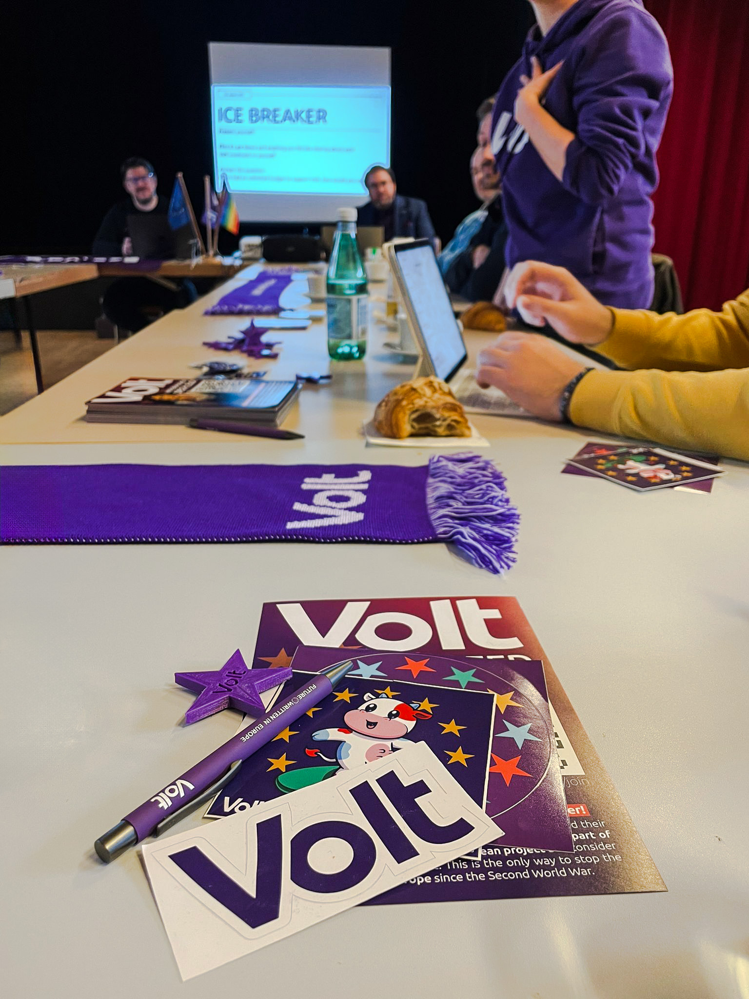 Volters are sitting at a table. In the foreground, you can see various Volters merchandising items on the table, such as stickers, a pen and a table magnet.