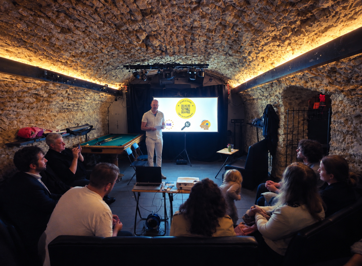 People sitting in a small room watching a screen, one person holding a speech